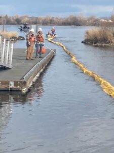 2 people stand on a dock while boom is in the water in the background there is a boat with people dragging the boom